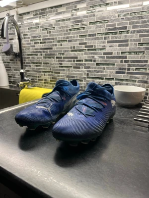 Puma future 7 fotbollskor - Säljer ett par blåa Puma fotbollsskor med vita detaljer och klassisk logga på sidan. Skorna har snörning, låg profil och greppvänliga dobbar för spel på gräs. Ovandelen är tillverkad i ett syntetmaterial som ger bra bollkontroll och komfort. Köpte den för 1099kr från stadium. Den är mycket bra skick, den är bara lite smutsig, går att tvätta