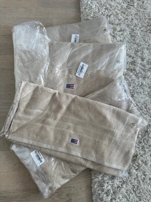 Beige handduk Lexington bomull - Säljer ett supersnyggt set från Lexington: ✨ 2 st badlakan, stora handdukar (tror 100x150 cm, kan öppna förpackning och dubbelkolla om så önskas) ✨ 2 st handdukar, mindre (tror 50x70 cm, kan öppna förpackning och dubbelkolla om så önskas) idealiska för händer/ansikte/gäst Alla i fin, neutral beige/sand-färg. Tror färgen ”Moonbeam”.  •  Original-serien från Lexington (kammad bomull, 600 g/m² – tjocka, absorberande och blir bara mjukare med tvätt) •  Helt nya – fortfarande i originalplast med etik