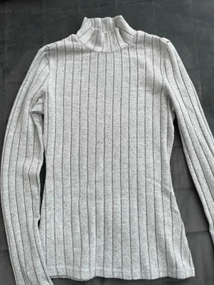 Ljusgrå ribbad polotröja - Säljer en ljusgrå ribbad polotröja med lång ärm. Tröjan har ett smalt, figurnära snitt och är tillverkad i ett mjukt, stretchigt material som känns skönt mot huden. Super bra för dig som letar efter en trendig och stily look. Passar jättefint med väst eller kofta över, annars med jeans, kostymbyxor eller kjol. 