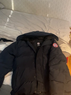 Mörk blå dunjacka från Canada Goose macmillan  - Säljer en mörkblå dunjacka från Canada Goose med klassisk logotyp på ärmen. Jackan har huva, dragkedja och knappar framtill samt två stora fickor. Perfekt för kalla vinterdagar och riktigt varm tack vare dunfyllningen.