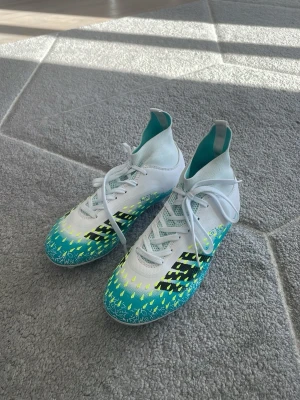 Adidas fotbollsskor - Säljer ett par snygga Adidas fotbollsskor med högt skaft och snörning. Skorna är vita med turkosa och gula detaljer samt svarta mönster på sidorna. Yttermaterialet är syntet och sulan har dobbar för bra grepp på planen. Perfekta för dig som vill sticka ut på fotbollsplanen! Storlek 38 aldrig använda.