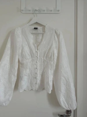 Vit broderad blus med puffärm Gina tricot  - Söt vit blus med broderade blommor, puffiga långärmar och knappar framtill. Blusen har v-ringning och lätt figursydd passform. Materialet är tunt och luftigt, perfekt för en fräsch och romantisk look.