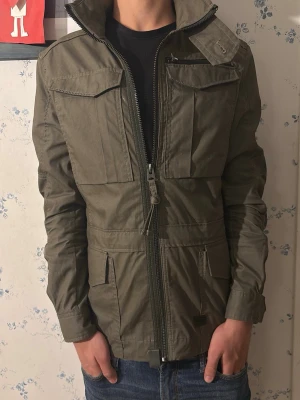 G-Star RAW Olivgrön fieldjacka  - Snygg olivgrön jacka från G-Star RAW med flera fickor framtill, snedställd dragkedja och klassisk krage. Jackan har en modern, rak passform . Perfekt för dig som gillar streetwear och funktionella detaljer. Passar allt från 170-180 isch 