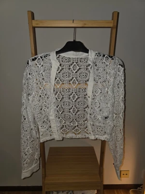 Vit spetskofta med knappar - Säljer en vit, långärmad kofta i spets med detaljerat virkat mönster och genomskinliga partier. Koftan har knappar framtill och ribbade kanter. Perfekt för att lyfta en outfit med en romantisk touch.