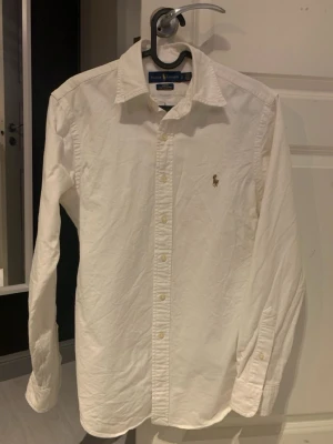 Ralph lauren skjorta  - Klassisk vit skjorta från Ralph Lauren med broderad logga på bröstet. Skjortan har lång ärm, knappar framtill och en snygg krage. Tillverkad i mjuk bomull som känns skön mot huden. Perfekt till jeans eller chinos för en clean look.