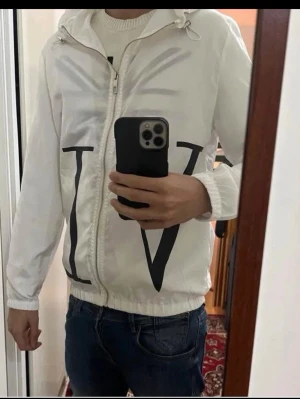 Vit vindjacka från Valentino - Snygg vit vindjacka från Valentino med stora svarta bokstäver tryckta fram och bak. Jackan har huva med dragsko, dragkedja framtill och elastiska muddar vid ärmar och midja. Tillverkad i lätt och glansigt material, perfekt för en trendig streetwear-look. Om vi möts upp i Stockholm kan jag gå ner i pris