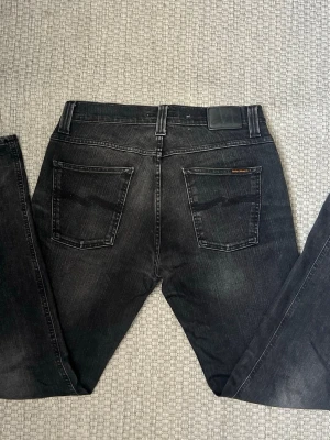 Svarta jeans från Nudie Jeans - Säljer ett par svarta jeans från Nudie Jeans med klassisk femficksdesign och raka ben. Jeansen har diskreta slitningar och Nudie-loggan på bakfickan. Tillverkade i mjuk denim som sitter snyggt och bekvämt.