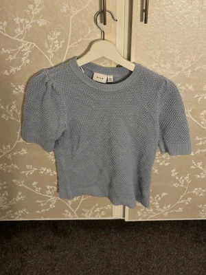 Ljusblå stickad topp från Vila - Supersöt ljusblå stickad topp från Vila med korta puffärmar och rund hals. Toppen har en ribbad kant vid ärmslut och nederkant, vilket ger en snygg och bekväm passform. Perfekt att styla med jeans eller kjol för en trendig look.