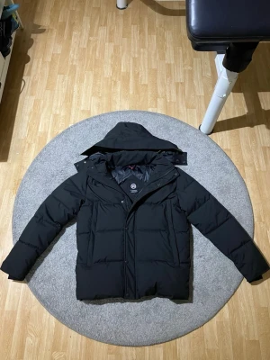 Svart dunjacka från Canada Goose - Säljer en svart dunjacka från Canada Goose med huva och diskret logga på ärmen. Jackan har en klassisk puffer-modell med dragkedja och knappar framtill. Perfekt för kalla vinterdagar och riktigt varm tack vare dunfyllningen. Passar också XL