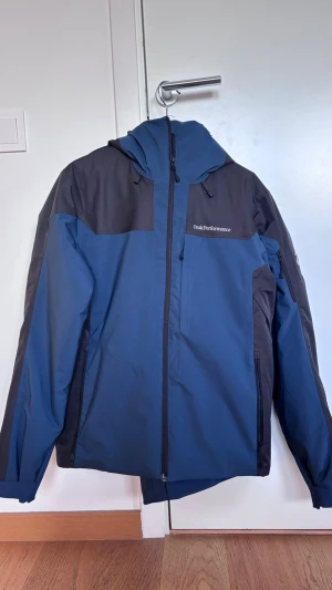 Blå och svart skidkläder Peak Performance - Snygg blå och svart jacka från Peak Performance med huva och dragkedja framtill. Använd endast en gång. Väldigt lätt och varm.  Köptes som unisex.  Modellen är Maroon Race Colour-Block.