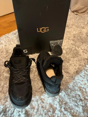 UGG LOWMEL - Snygga svarta sneakers från UGG med robust sula och skön innersida i mjukt foder. Skorna har textil- och mockadetaljer, svarta skosnören och UGG-logga på plösen. Perfekta för dig som vill ha både stil och komfort.