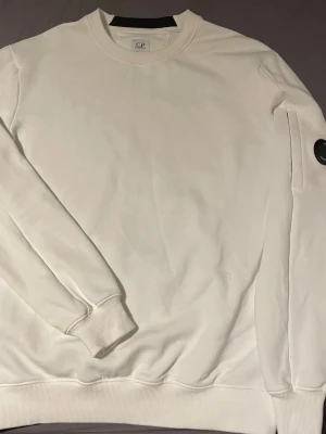 Vit sweatshirt från C.P. Company - Säljer en stilren vit sweatshirt från C.P. Company med rund halsringning och ribbade muddar. Tröjan har en ikonisk svart linsdetalj på ena ärmen och är tillverkad i mjukt bomullsmaterial. Perfekt för en clean och modern look.