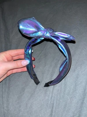Diadem - Säljer ett diadem i skimrande material med en stor knuten rosett på toppen. Färgen skiftar mellan blått, lila och turkos beroende på ljuset. Diademet har en bred form och ger en riktigt trendig vibe till din look. Inga defekter