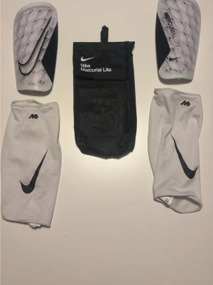 Nike Mercurial Lite benskydd set - Säljer ett komplett set Nike Mercurial Lite benskydd i vitt och svart. Setet inkluderar två benskydd med bikakemönster, två vita strumpor med svart swoosh och en svart meshväska med Nike-logga. Perfekt för fotbollsträning eller match.