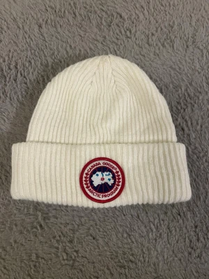 Vit ribbstickad mössa Canada Goose - Snygg vit ribbstickad mössa EJ från Canada Goose med klassisk rund logotyp framtill. Mössan har uppvikt kant och är tillverkad i mjukt material som håller dig varm. Perfekt accessoar för kalla dagar och enkel att matcha med olika outfits.