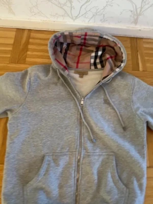 Grå zip-hoodie från Burberry - Snygg grå hoodie med dragkedja från Burberry. Huvan är fodrad med det klassiska Burberry-rutiga mönstret i beige, svart och rött. Hoodien har fickor framtill och justerbar dragsko i huvan. Perfekt för en avslappnad och trendig look.