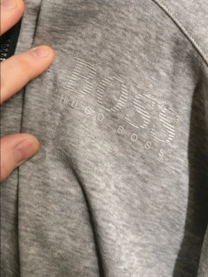 Grå hoodie från Hugo Boss Helg REA NU! - Snygg grå hoodie från Hugo Boss med dragkedja och ribbad krage. Stort BOSS-tryck på bröstet och detaljerad dragkedja med logga. Mjuk bomullskänsla och klassisk passform, perfekt för en stilren och avslappnad look.