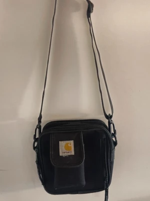 Svart axelväska från Carhartt - Säljer en svart axelväska från Carhartt med justerbar rem, flera fack och dragkedjor. Väskan har Carhartt-logga på framsidan och är tillverkad i 100% polyester. Perfekt för att bära det viktigaste och har en stilren, fyrkantig form.