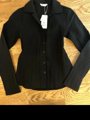 Svart figursydd blus  - Superfin svart blus med lätt strukturerat tyg som ger ett exklusivt och modernt intryck. Figursydd passform som sitter riktigt snyggt på kroppen. Klassisk krage och knäppning framtill – perfekt både till skolan, middagar eller när du vill ha en mer uppklädd vardagslook.