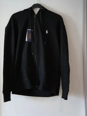 Svart hoodie från Polo Ralph Lauren - Snygg svart hoodie med dragkedja från Polo Ralph Lauren. Klassisk design med huva och justerbara snören, samt vit broderad logga på bröstet. Tillverkad i mjukt material som känns skönt mot huden. Perfekt för en avslappnad och stilren look. Utan prislappen