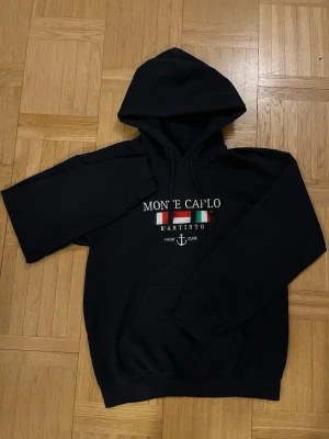 Marinblå hoodie Monte Carlo Kartisto - Svart hoodie från Kartisto med trycket 'Monte Carlo' och färgglada flaggor på bröstet. Klassisk känguruficka framtill, dragsko i huvan och ribbade muddar. Perfekt för en avslappnad och sportig look.