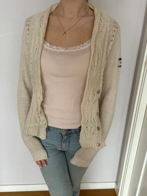 Beige stickad kofta med knytband  - Beige stickad kofta från Odd Molly med fina virkade detaljer och knytband i midjan. Koftan har långa ärmar, öppen front och dekorativa knappar. Perfekt för lager på lager och ger en avslappnad men ändå söt look. 💓