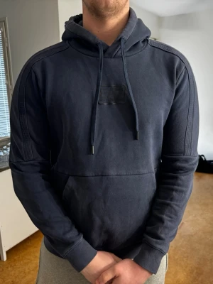 Emporio Armani hoodie - Snygg mörkblå hoodie från EA7 Emporio Armani med broderad logga på bröstet. Tröjan har en stor huva med snörning, ribbade muddar och en ficka framtill. Storlek L, nypris 1200 kr  Det är bara att skriva om ni vill ha mer frågor eller bilder!