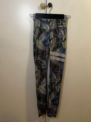 Mönstrade leggings STRONGER - Snygga leggings från STRONGER med tropiskt bladmönster i grönt, blått och brunt. Hög midja och smal passform, samt dekorativa vita ränder på ena benet. Perfekta för träning eller chill. Materialet är stretchigt och bekvämt.