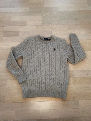 Grå kabelstickad tröja Polo Ralph Lauren - Kabelstickad grå tröja från Polo Ralph Lauren med rund halsringning och ribbade muddar. Klassisk svart broderad logga på bröstet. Tillverkad i mjukt material som ger en skön och avslappnad look. Storleken är snarare sagt small, även fast att den är medium.