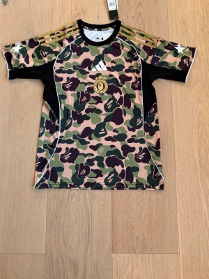 Adidas x BAPE camo t-shirt - Säljer en Adidas x BAPE t-shirt med ikoniskt grönt, beige och brunt camomönster. Tröjan har svarta sidopaneler, guldfärgade ränder på axlarna och tryckt BAPE-logga på bröstet. Tillverkad i ett lätt och funktionellt material, perfekt för sport eller streetwear.