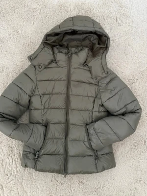 Olivgrön pufferjacka med huva - Snygg olivgrön pufferjacka med avtagbar huva och dragkedja framtill. Jackan har två praktiska fickor med dragkedja och quiltad design. Perfekt för kalla dagar och ger en stilren look. Materialet är glansigt och lätt vadderat för extra värme.