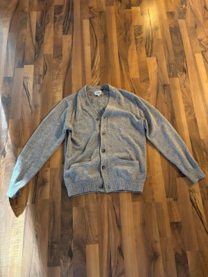 Grå cardigan med knappar  - En enkel grå kofta med v-ringning och fem knappar framtill. Koftan har två fickor fram och ribbade muddar vid ärmslut och nederkant. Perfekt att slänga över en t-shirt för en avslappnad look eller över en tunnare tröja. Tröjan är i perfekt skick! Passar S/M