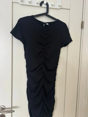 Svart kortklänning med rynk - Snygg svart kortklänning med rynkdetaljer framtill och korta ärmar. Klänningen har en figurnära passform och är tillverkad i ett mjukt, stretchigt material. Perfekt för dig som gillar en enkel men trendig look.