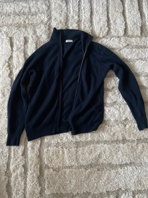 Mörkblå soft goat fullzip tröja - Mörkblå ziptröja med hög krage och hel dragkedja framtill. Tröjan har ribbade muddar och snygga mockalappar på armbågarna och 100% Kashmir. Nypris: 3200kr        