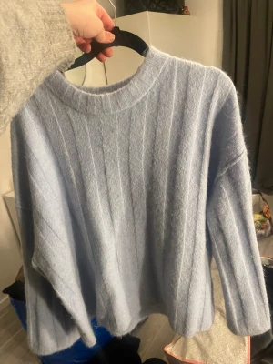 Ljusblå oversized stickad tröja - Mysig ljusblå stickad tröja med breda ribbade ränder och avslappnad passform. Tröjan har rund halsringning och långa ärmar, perfekt för lager på lager. Materialet ser fluffigt och mjukt ut, vilket ger en skön känsla.