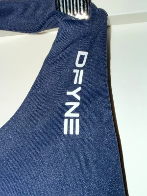 Mörkblå sporttopp från DFYNE - Snygg mörkblå sporttopp från DFYNE med breda axelband och vit logga på axeln. Tillverkad i stretchigt funktionsmaterial som passar perfekt för träning och gym. Enkel, stilren design med tight passform. Helt nya!