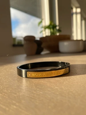 Svart armband från MichaelKors - Stilrent svart armband från MichaelKors med en guldfärgad metallplatta där det står 'MICHAELKORS EST. 1981'. Armbandet har en modern och minimalistisk design som passar perfekt till din vardagsstil. Tillverkat i metall med en blank finish.