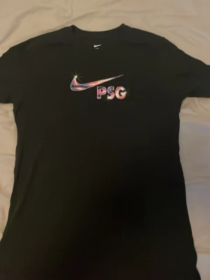 Svart PSG x Nike t-shirt - Snygg svart t-shirt från Nike med PSG-logga och färgglad swoosh på bröstet. Klassisk rund hals och korta ärmar. Perfekt för dig som gillar sportig stil och vill representera Paris Saint-Germain. Materialet känns mjukt och skönt mot huden.
