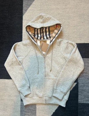Grå zip hoodie från Burberry - Säljer en grå zip hoodie från Burberry med klassiskt rutigt foder i huvan och dragsko. Hoodien har ribbade muddar, fickor framtill och är tillverkad i mjuk bomullsblandning. Perfekt för en avslappnad och stilren look.
