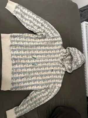 Mönstrad hoodie från Lacoste LIVE - Säljer en loose fit hoodie från Lacoste LIVE i unisexmodell. Hoodien är vit med grönt geometriskt mönster och har ribbade muddar vid ärmslut och nederkant. Utrustad med huva och dragsko. Perfekt för dig som gillar streetwear och unik design.