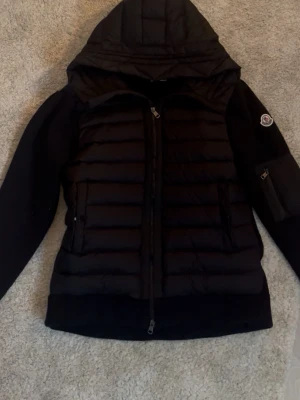 Svart cardigan från Moncler - JAG TAR BYTEN. Snygg svart cardigan från Moncler med huva och quiltad front. Jackan har dragkedja framtill, två sidofickor med dragkedja och en extra ficka på ärmen med Moncler-logga. Materialet är en mix av glansigt och matt tyg för en cool kontrast. Den kommer med alla taggar, lappar och påsar. Har aldrig använt