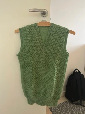 Grön stickad väst med v-ringning - Snygg grön stickad väst med v-ringning och grovt mönster framtill. Västen är ärmlös och har ribbade kanter vid hals, ärmhål och nederkant. Perfekt att bära över en skjorta eller t-shirt för en trendig lager-på-lager-look.