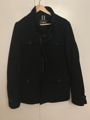Svart jacka från Dressmann - Stilren svart jacka från Dressmann med klassisk krage och fyra fickor framtill. Jackan har knappar och dragkedja. Normal i storleken och sparsamt använd, hängt i garderoben ett långt tag.