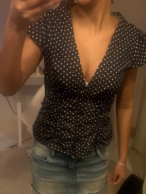 Svart prickig blus med v-ringning - Snygg svart blus med vita prickar och djup v-ringning. Modellen är figurnära med knappar framtill och korta ärmar. Perfekt till jeans eller kjol för en trendig look. Materialet känns lätt och bekvämt.