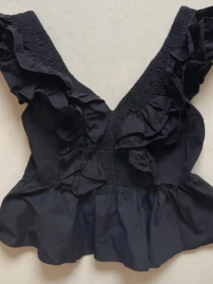 Svart volangtopp med dragkedja - Supersnygg svart topp med breda axelband och volangdetaljer framtill och baktill. Toppen har smock över axlarna och en markerad midja med volang nedtill. Dragkedja i ryggen för enkel påklädning. Perfekt för dig som vill sticka ut lite extra.