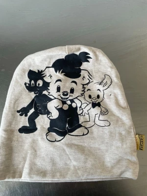 Bamse mössa med tryck i nyskick - Ljusgrå mössa med stort svart tryck av Bamse, Lille Skutt och Skalman på framsidan. Tillverkad i mjukt bomullsmaterial och har en gul Bamse-etikett i sidan. Perfekt för dig som gillar klassiska seriekaraktärer.