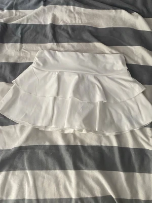 Vit volangkjol med shorts från SHEIN - Säljer en vit kortkjol från SHEIN Teen med dubbla volanger. Kjolen har en enkel och stilren design, den har också vita shorts under. Tillverkad med lite stretch för extra komfort.