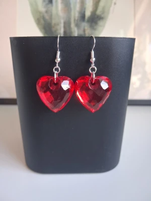 Röda hjärtformade örhängen - Säljer ett par örhängen med stora, röda hjärtan i facetterad plast som ger en glittrig effekt. Krokarna är i metallfärg och hjärtana har en tydlig poppig look. Perfekta för dig som vill sticka ut med en färgstark accessoar. Mått 22x22x11 ca 3cm långa. Öronkroken är nickelsäker och aldrig använda 