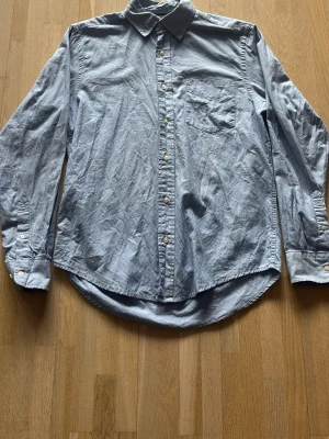 Blå skjorta från Tommy hilfiger - Klassisk blå skjorta i bomull med lång ärm och bröstficka. Skjortan har knappar framtill och på manschetterna samt en snygg krage. Perfekt för en clean och enkel stil.