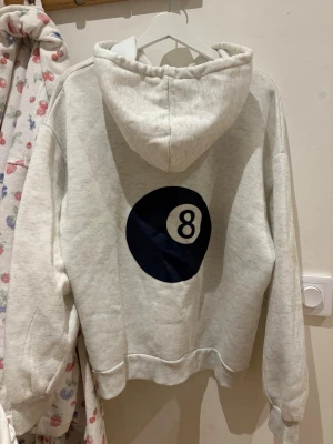 Vit hoodie med 8-ball tryck - Säljer en oversized vit hoodie med stor svart 8-ball print på ryggen och en mindre på bröstet. Hoodien har huva med dragsko och en stor magficka. Tillverkad i mjukt sweatshirtmaterial, perfekt för en chill och avslappnad stil.